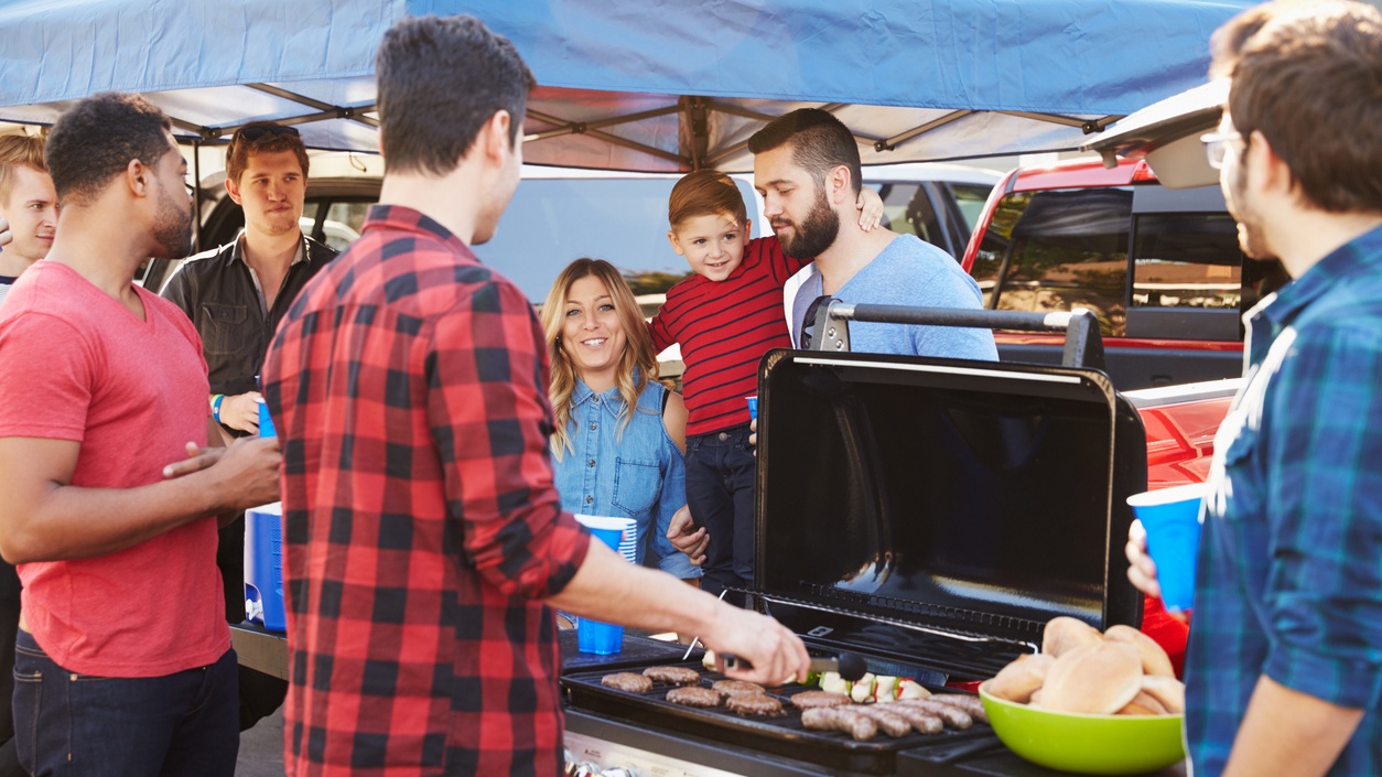 Adults and children crowd around a grill at a tailgate.