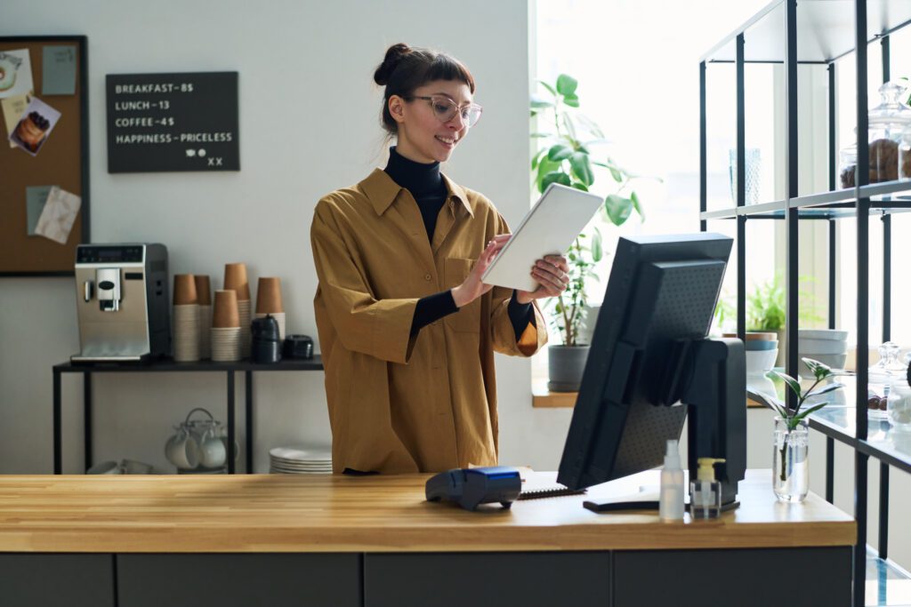 Small Business owners in Indiana appreciate the power of Comcast Business and its network to keep mobile devices online with fast, secure and reliable internet. IMAGE: Happy young clerk of cafe using tablet while standing by counter desk in front of computer monitor and taking online orders or counting cost